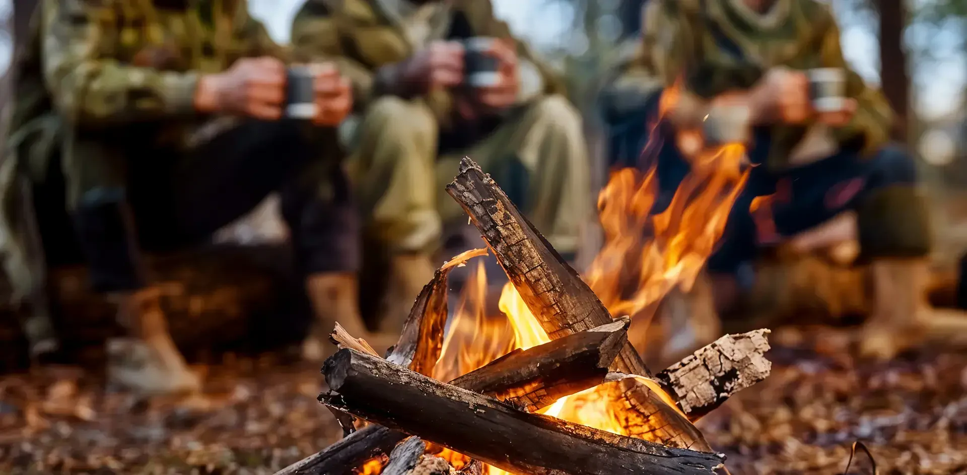 Campfire with orange flames, logs, and foliage; people enjoying drinks nearby.