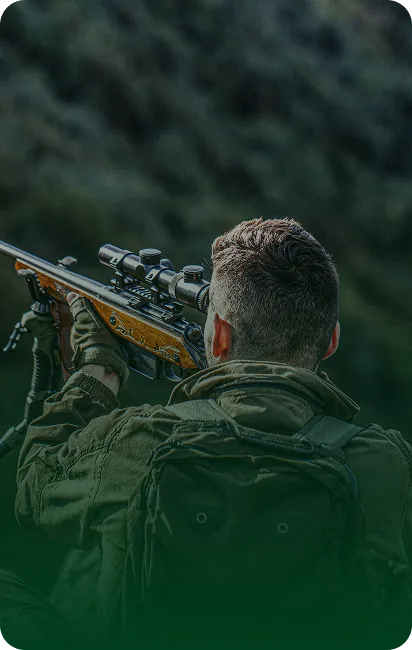 Soldier in tactical gear aiming a sniper rifle in a rugged landscape.