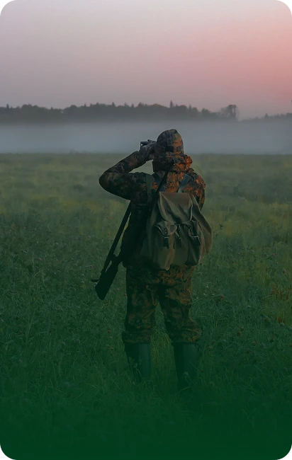 Hunter in camouflage at dawn with a camera and rifle, standing in a misty field.
